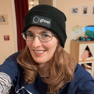 A photo of Cathy. She's wearing a black beaning that says Ghost. Cathy is a white woman with glasses, blue eyes, brown hair, and freckles.